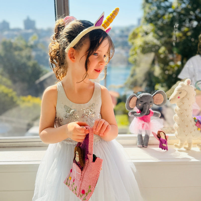 Girl wearing a unicorn headband holding a handmade bag, surrounded by plush toys, celebrating with a bright smile.