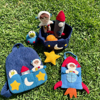 A set of handmade felt space-themed pouches and small plush toys arranged on green grass. The set includes a blue astronaut window pouch, a blue rocket-shaped pouch with a red nose and orange tail, a dark blue moon basket with a small astronaut plush, a small blue frog plush, and a yellow star.