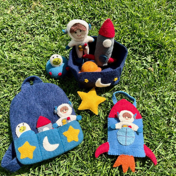A set of handmade felt space-themed pouches and small plush toys arranged on green grass. The set includes a blue astronaut window pouch, a blue rocket-shaped pouch with a red nose and orange tail, a dark blue moon basket with a small astronaut plush, a small blue frog plush, and a yellow star.