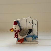 Christmas Seagull with chip decoration featuring a seagull in a Santa hat holding a chip next to a wooden fence.